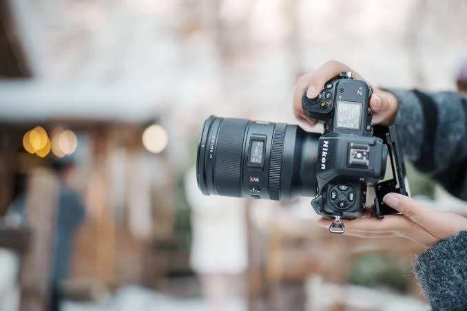 Фотограф у зимовому одязі тримає камеру Nikon Z8 з об'єктивом Viltrox LAB на фоні розмитого зимового пейзажу з боке від вогників.